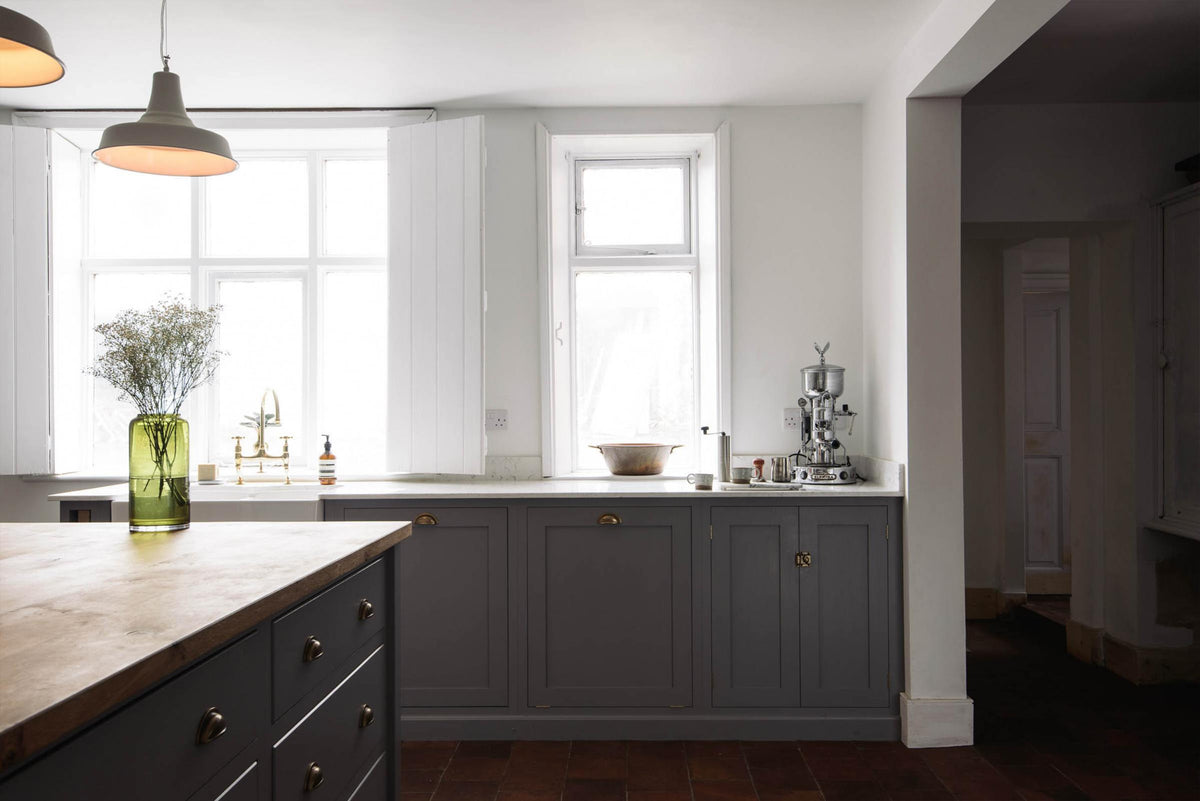 Bright light from the open windows highlights a simple, modern Shaker kitchen in a historic Cheshire townhouse. It features a cooker run, Silestone sink run with a Belfast sink and aged brass Ionian tap, plus a large island with a solid oak worktop and breakfast nook. Painted in muted cool grey with white walls and warm terracotta tile flooring.