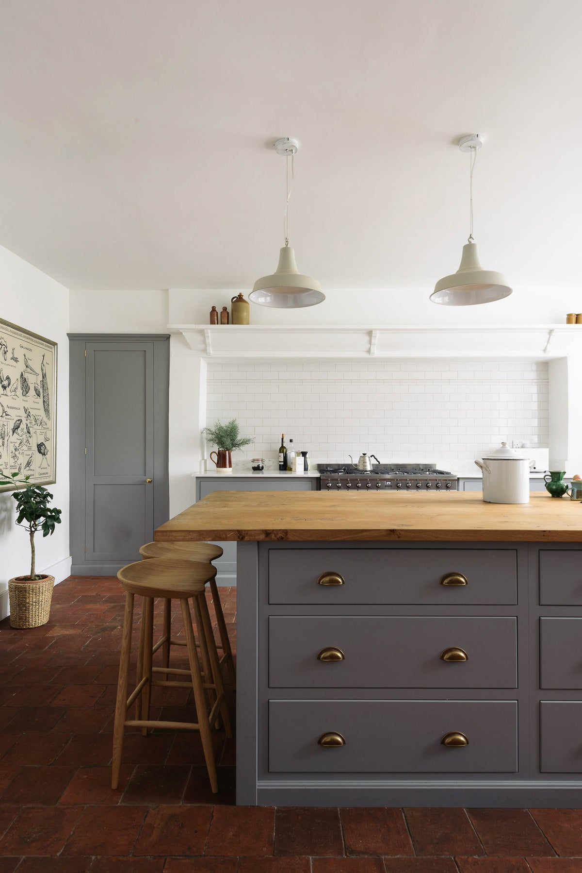 A close view at the solid oak bum stools tucked into the breakfast nook of a simple, modern Shaker kitchen island in a historic Cheshire townhouse. The kitchen features a cooker run with white porcelain splashback tiles, muted cool grey cupboards, white walls, and warm terracotta flooring.