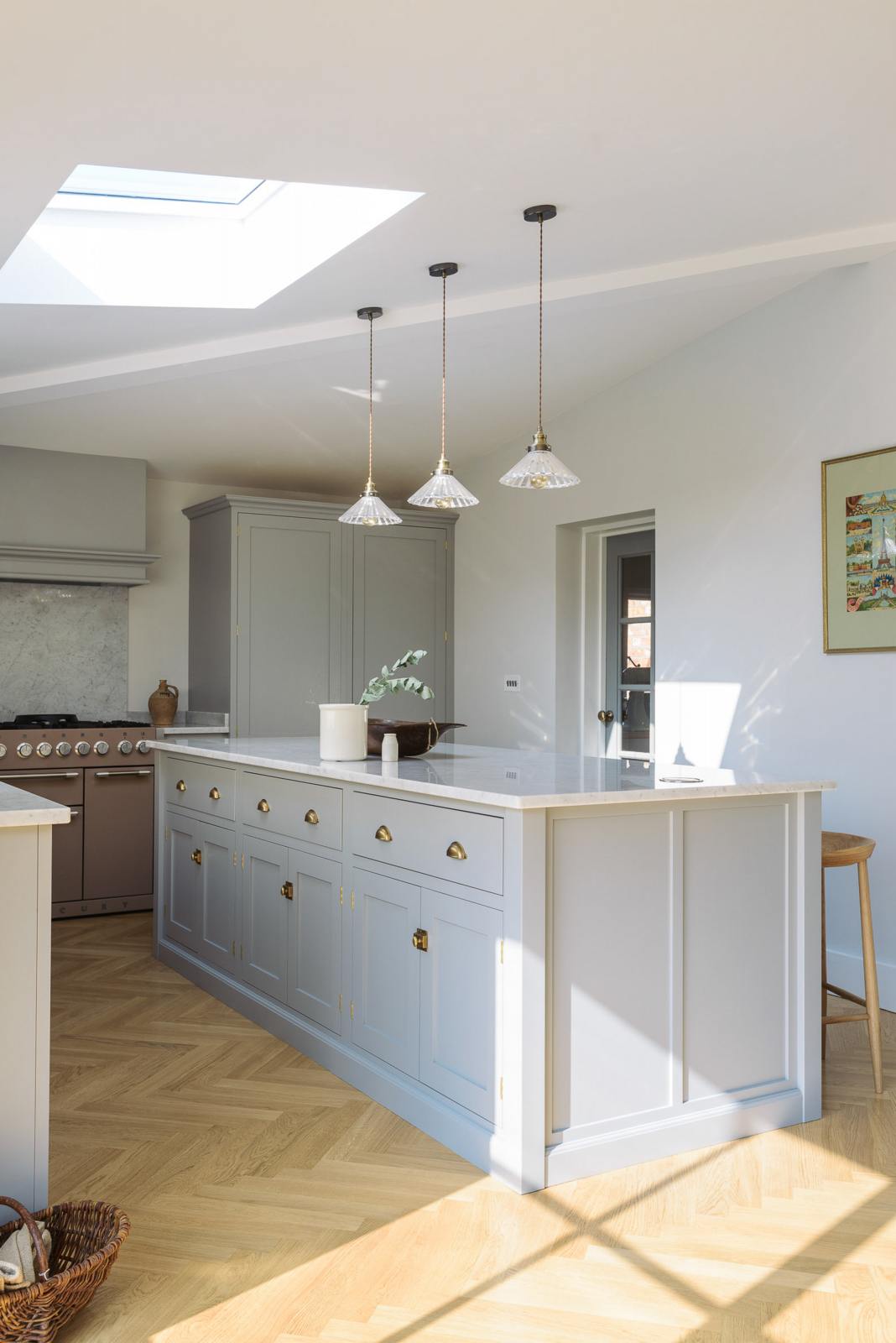 An oblique view of a spacious and contemporary stone grey Shaker kitchen features an L-shaped sink and cooker run with a Carrara worktop and splashback, a canopy, a bank of full-height cabinets, and a kitchen island. Elegantly decorated with glass-frilled pendant lights, artisan ceramics, and a deVOL bum stool, it creates a cool and inviting space.