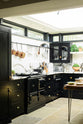 An angled view of a Painter’s Black shake kitchen with an L-shaped layout featuring a cooker and a sink. The worktop and splashback are crafted from San Simone quartzite. The design includes an overhead glazed cupboard and a tall storage cupboard, along with an aged brass hanging rail. The space is illuminated by a glass skylight, enhancing its overall brightness.