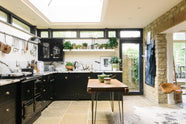 A beautifully renovated kitchen features a Painter’s Black shaker style with an L-shaped layout that includes a cooker and sink run. The worktop and splashback are made from San Simone quartzite, complemented by an overhead glazed cupboard and a tall storage cupboard. A glass skylight brightens the modern space while honouring the historical character of the building by retaining the original brick wall.