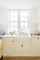 A close-up view of a small, white linen shaker kitchen sink run featuring a double Belfast sink, an aged brass Ionian tap, and a Carrara marble worktop, overlooking a glass window with a cityscape view.