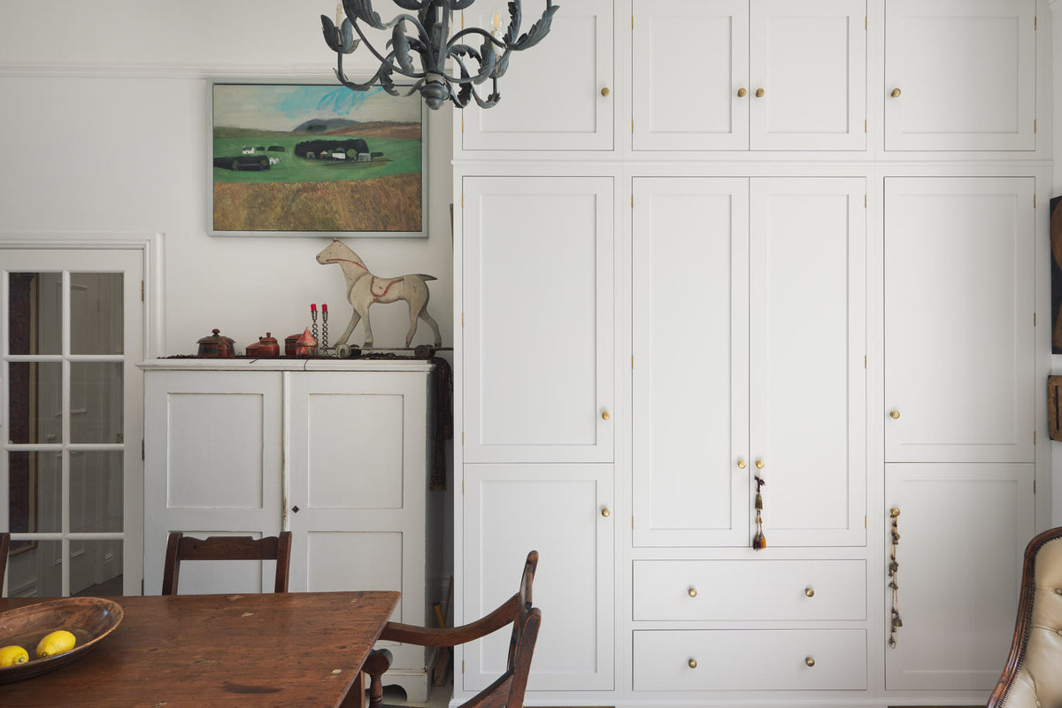 A quiet corner of a tall yet simple Shaker kitchen featuring a small bank of larder. Lovingly decorated with antique furniture, a vibrant red decorative rug, a variety of handmade charms, and a vintage white armchair.