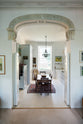 Through a grand decorative archway is a tall yet simple Shaker kitchen featuring a sink and cooker run with a Carrara marble worktop, shelves, and a small bank of larder. Lovingly decorated with antique furniture, a vibrant red decorative rug, a variety of chic and curated paintings, and a vintage metal chandelier.