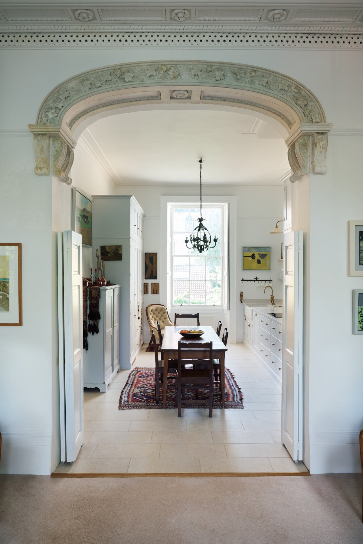 Through a grand decorative archway is a tall yet simple Shaker kitchen featuring a sink and cooker run with a Carrara marble worktop, shelves, and a small bank of larder. Lovingly decorated with antique furniture, a vibrant red decorative rug, a variety of chic and curated paintings, and a vintage metal chandelier.