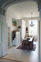 An oblique view of a tall yet simple Shaker kitchen featuring a sink and cooker run with a Carrara marble worktop, shelves, and a small bank of larder. Lovingly decorated with antique furniture, a vibrant red decorative rug, a variety of chic and curated paintings and artworks, and a vintage metal chandelier.