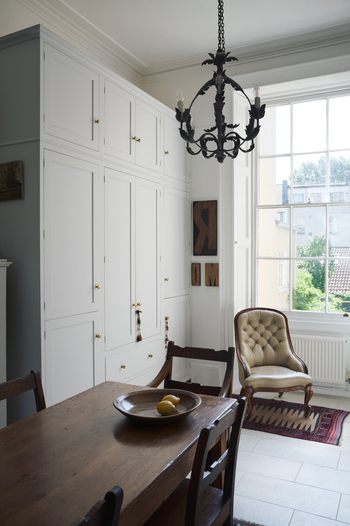 A close, oblique view of a tall yet simple Shaker kitchen featuring a sink and cooker run with a Carrara marble worktop, shelves, and a small bank of larder. Lovingly decorated with antique furniture, a vibrant red decorative rug, a variety of chic and curated paintings and artworks, and a vintage metal chandelier.