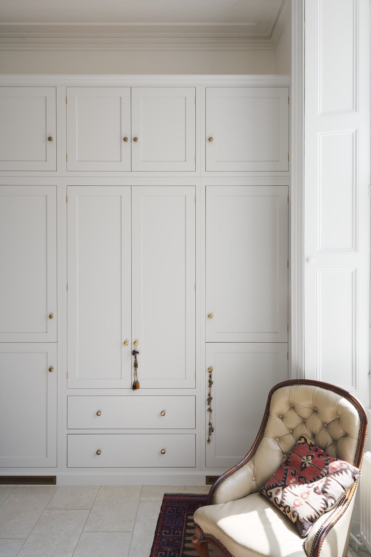 A quiet corner of a tall yet simple Shaker kitchen featuring a small bank of larder. Lovingly decorated with antique furniture, a vibrant red decorative rug, a variety of handmade charms, and a vintage white armchair.