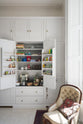 A quiet corner of a tall yet simple Shaker kitchen featuring a small bank of larder and an open pantry cupboard. Lovingly decorated with everyday kitchen equipment, spice, books, antique furniture, a vibrant red decorative rug, a variety of handmade charms, and a vintage white armchair.