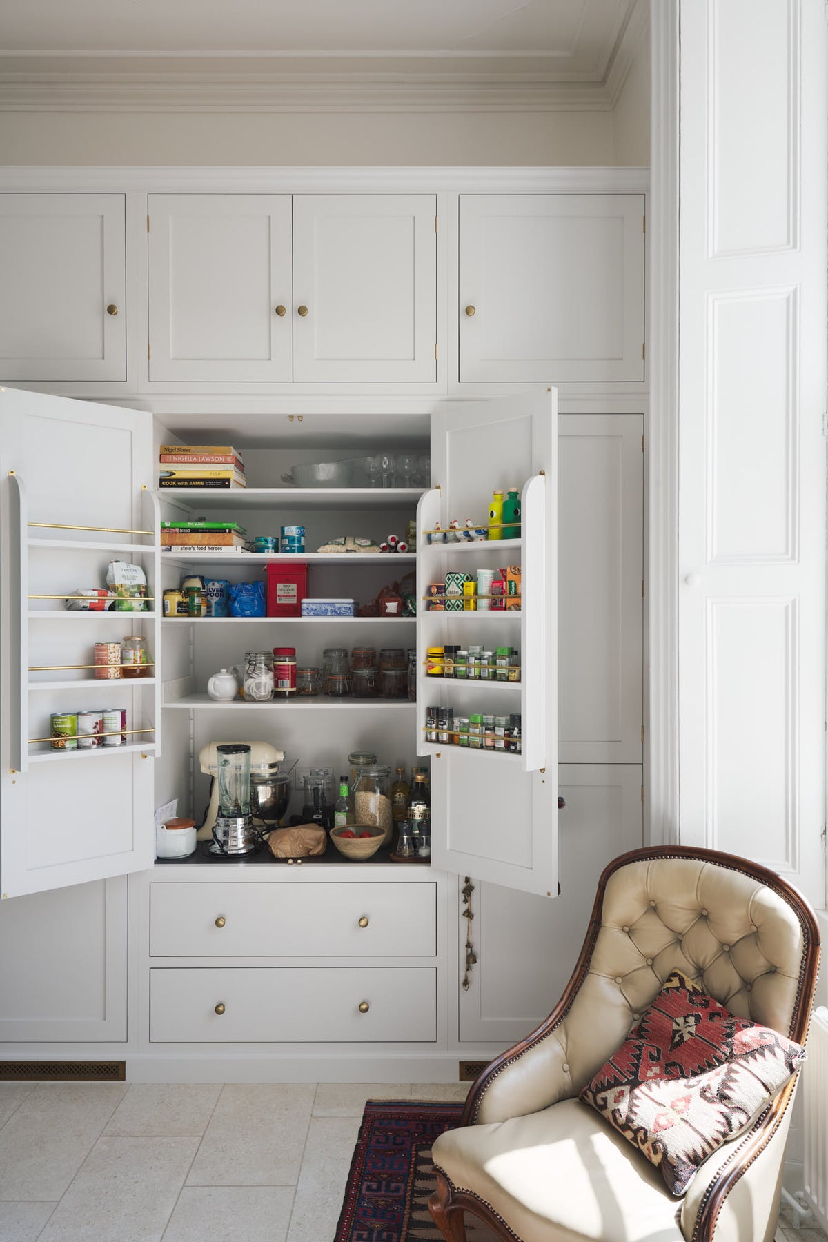 A quiet corner of a tall yet simple Shaker kitchen featuring a small bank of larder and an open pantry cupboard. Lovingly decorated with everyday kitchen equipment, spice, books, antique furniture, a vibrant red decorative rug, a variety of handmade charms, and a vintage white armchair.