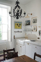 An oblique view of a quiet corner of a tall yet simple Shaker kitchen featuring a sink and cooker run with a Carrara marble worktop, shelves, an aged brass Ionian tap, and a Heirloom task light. Lovingly decorated with antique furniture, artisan ceramics, a variety of chic and curated paintings, and a vintage metal chandelier and hook rack.