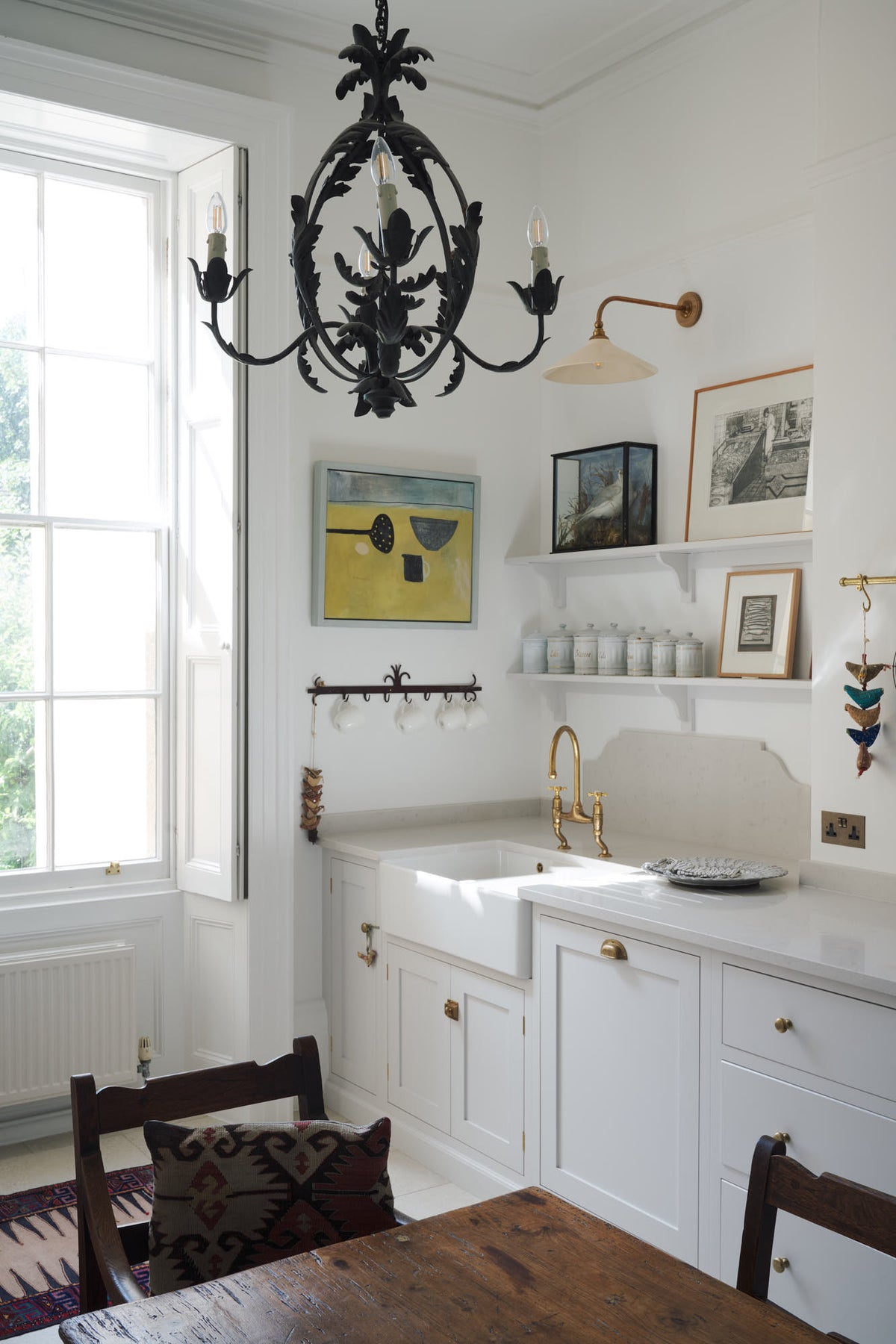 An oblique view of a quiet corner of a tall yet simple Shaker kitchen featuring a sink and cooker run with a Carrara marble worktop, shelves, an aged brass Ionian tap, and a Heirloom task light. Lovingly decorated with antique furniture, artisan ceramics, a variety of chic and curated paintings, and a vintage metal chandelier and hook rack.