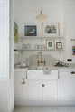 A quiet corner of a tall yet simple Shaker kitchen featuring a sink and cooker run with a Carrara marble worktop, shelves, an aged brass Ionian tap, and a Heirloom task light. Lovingly decorated with antique furniture, artisan ceramics, a variety of chic and curated paintings, and a vintage metal hook rack.