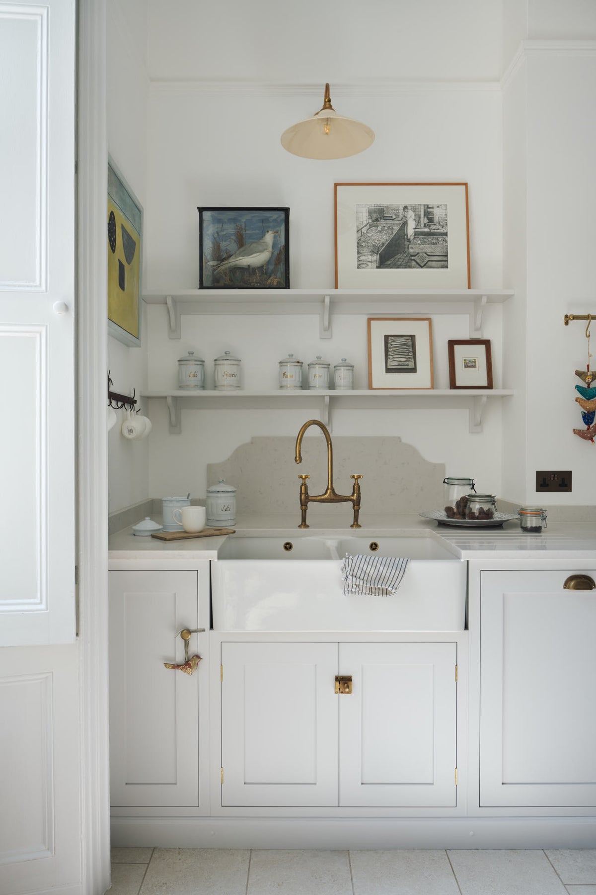 A quiet corner of a tall yet simple Shaker kitchen featuring a sink and cooker run with a Carrara marble worktop, shelves, an aged brass Ionian tap, and a Heirloom task light. Lovingly decorated with antique furniture, artisan ceramics, a variety of chic and curated paintings, and a vintage metal hook rack.