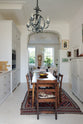 A tall yet simple Shaker kitchen featuring a sink and cooker run with a Carrara marble worktop, shelves, a small bank of larder, and a grand decorative archway towards the living room. Lovingly decorated with antique furniture, a vibrant red decorative rug, a variety of chic and curated paintings, and a vintage metal chandelier.