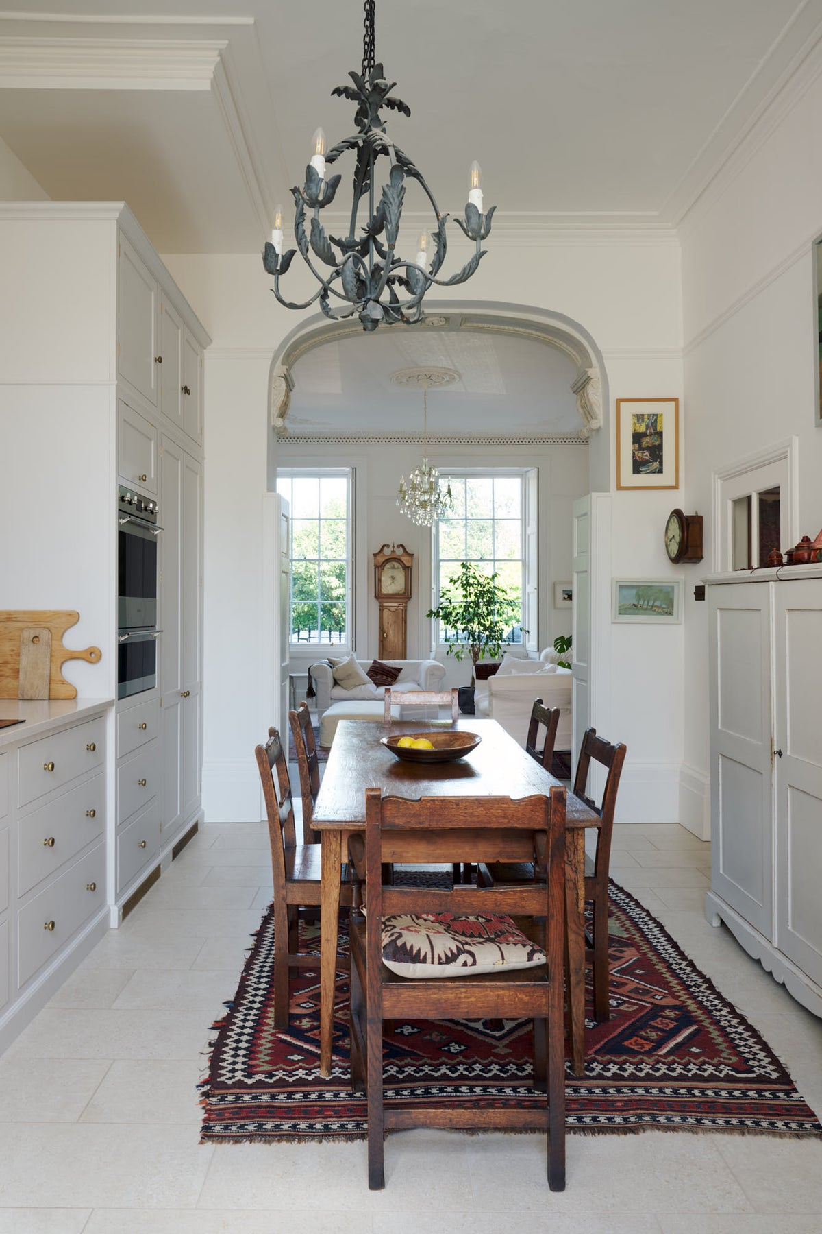 A tall yet simple Shaker kitchen featuring a sink and cooker run with a Carrara marble worktop, shelves, a small bank of larder, and a grand decorative archway towards the living room. Lovingly decorated with antique furniture, a vibrant red decorative rug, a variety of chic and curated paintings, and a vintage metal chandelier.