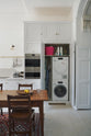 A practical corner of a tall yet simple Shaker kitchen featuring a sink and cooker run with a Carrara marble worktop, an oven cabinet, and an open appliance cupboard revealing a washing machine and dryer. Lovingly decorated with antique furniture, a vibrant red decorative rug, a variety of chic and curated paintings, and a vintage metal chandelier.