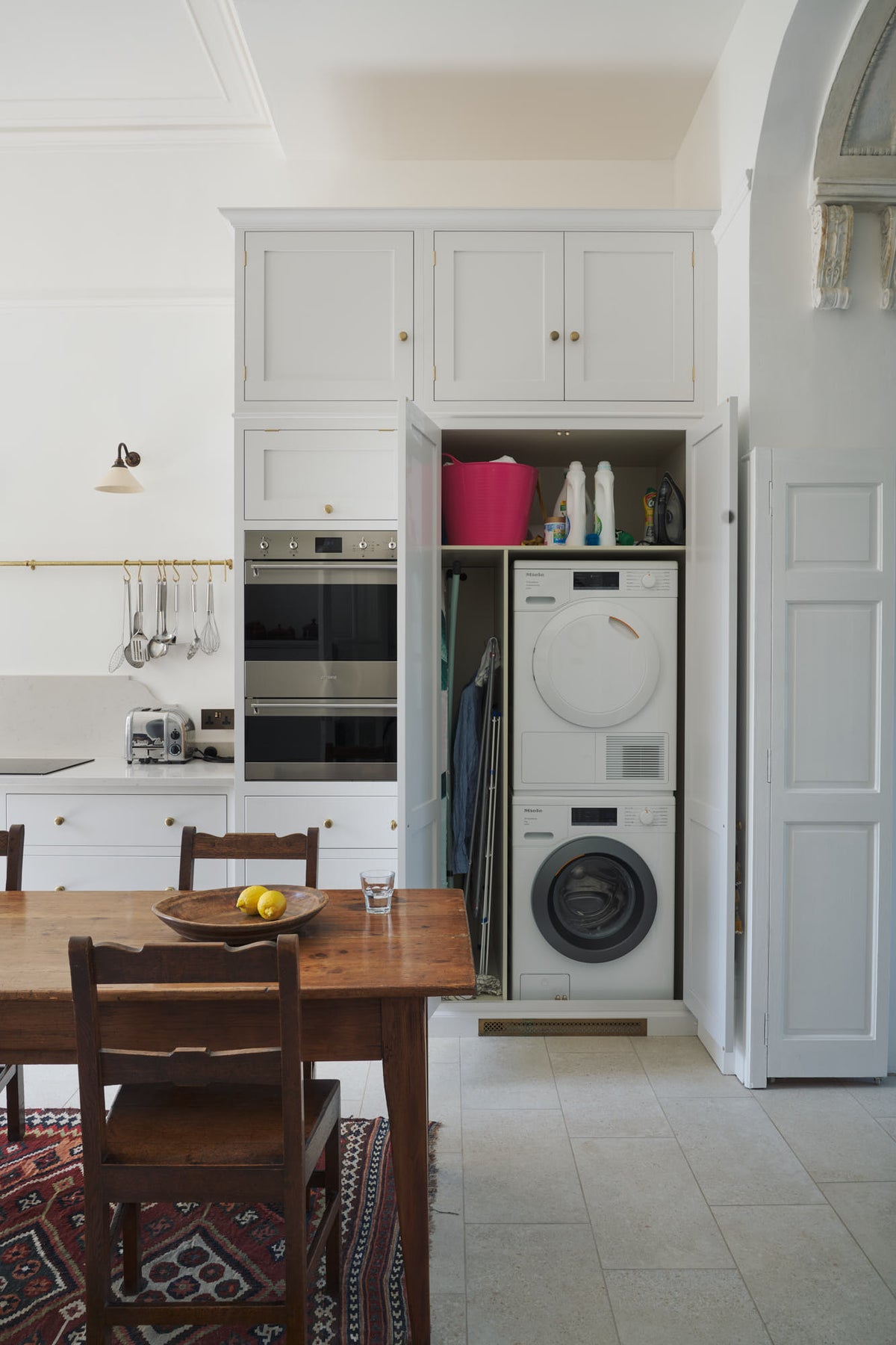 A practical corner of a tall yet simple Shaker kitchen featuring a sink and cooker run with a Carrara marble worktop, an oven cabinet, and an open appliance cupboard revealing a washing machine and dryer. Lovingly decorated with antique furniture, a vibrant red decorative rug, a variety of chic and curated paintings, and a vintage metal chandelier.