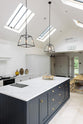An oblique view of a spacious and artisan Shaker kitchen features an off-white L-shaped sink run with a double Belfast sink and an aged brass Ionian tap, a pantry-blue kitchen island with a Carrara marble worktop, a canopy, and a bank of cupboards. Cleverly decorated with steel-framed hanging lights, vintage ceramics, and the original barn door serving as artistic displays, it creates a simple yet intriguing kitchen space.