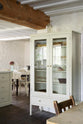 An angled shot of a small, free-standing cream cupboard, displaying assorted wine glasses, ceramics and a seashell. Part of the simple English farmhouse style kitchen in deVOL’s headquarters at Cotes Mill, with low, exposed wooden beams and worn wooden floorboards. Scattered is a collection of vintage items, including chairs, tables, and bric-a-brac, beautifully coordinated to complement the Mushroom Shaker kitchen.
