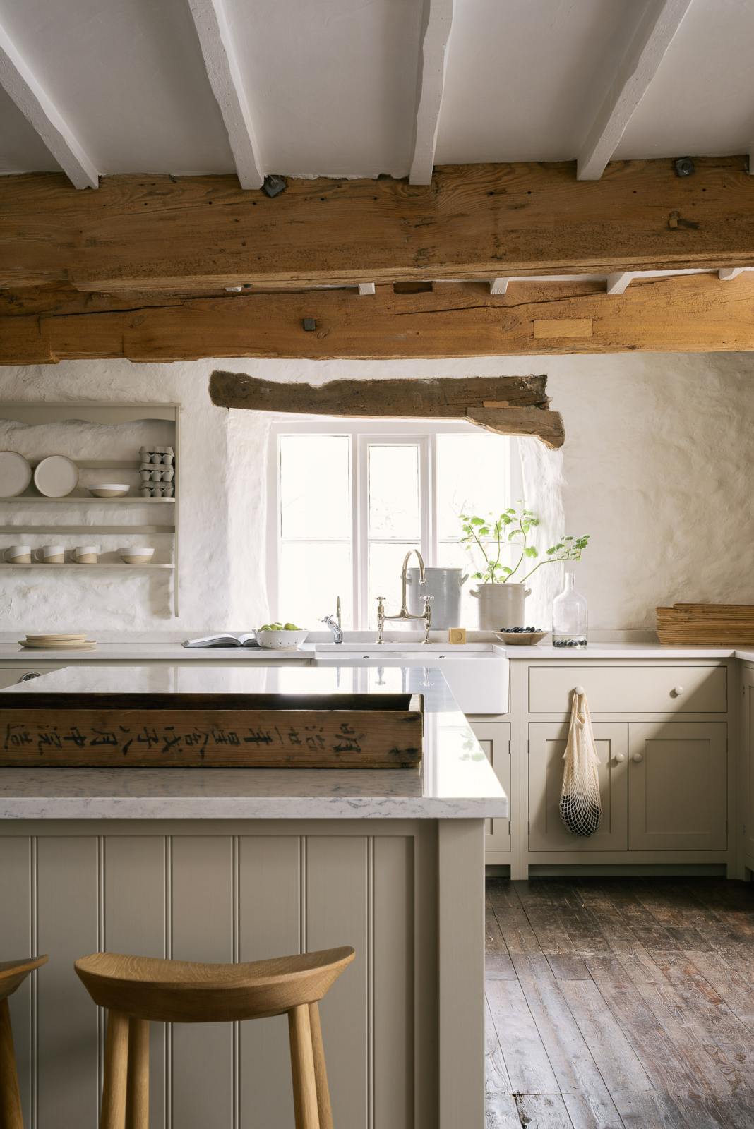 A simple English farmhouse kitchen in deVOL’s headquarters at Cotes Mill, with low, exposed wooden beams and worn wooden floorboards. The walls are textured and wobbly and painted in a light, off-white shade. The Shaker sink run, island and cooker run are all painted in Mushroom, a soft beige colour that feels timeless and classic.