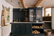A sample pantry blue shaker kitchen’s sink run features a shelf, countertop glazed cupboards, and a mix of closed and open base cabinets. The Carrara marble worktop holds a copper single sink with aged brass Mayan taps. Wooden and metal accents—including firewood, a metal bin, and a wooden toolbox—add rustic charm.