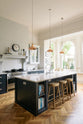 The pantry blue shaker kitchen features creamy Silestone worktops and off-white walls with two pale wall cupboards flanking the chimney breast. The central island has a sink on one side and a breakfast bar with wooden stools on the other, illuminated by three long brass lights above the prep table.