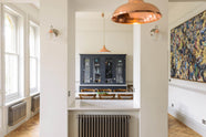 Two columns stand centre room, framing the dark navy blue cupboard. A rustic table with vintage wooden chairs sits before it, lit by a hanging light. The space features herringbone wooden floors and off-white walls.
