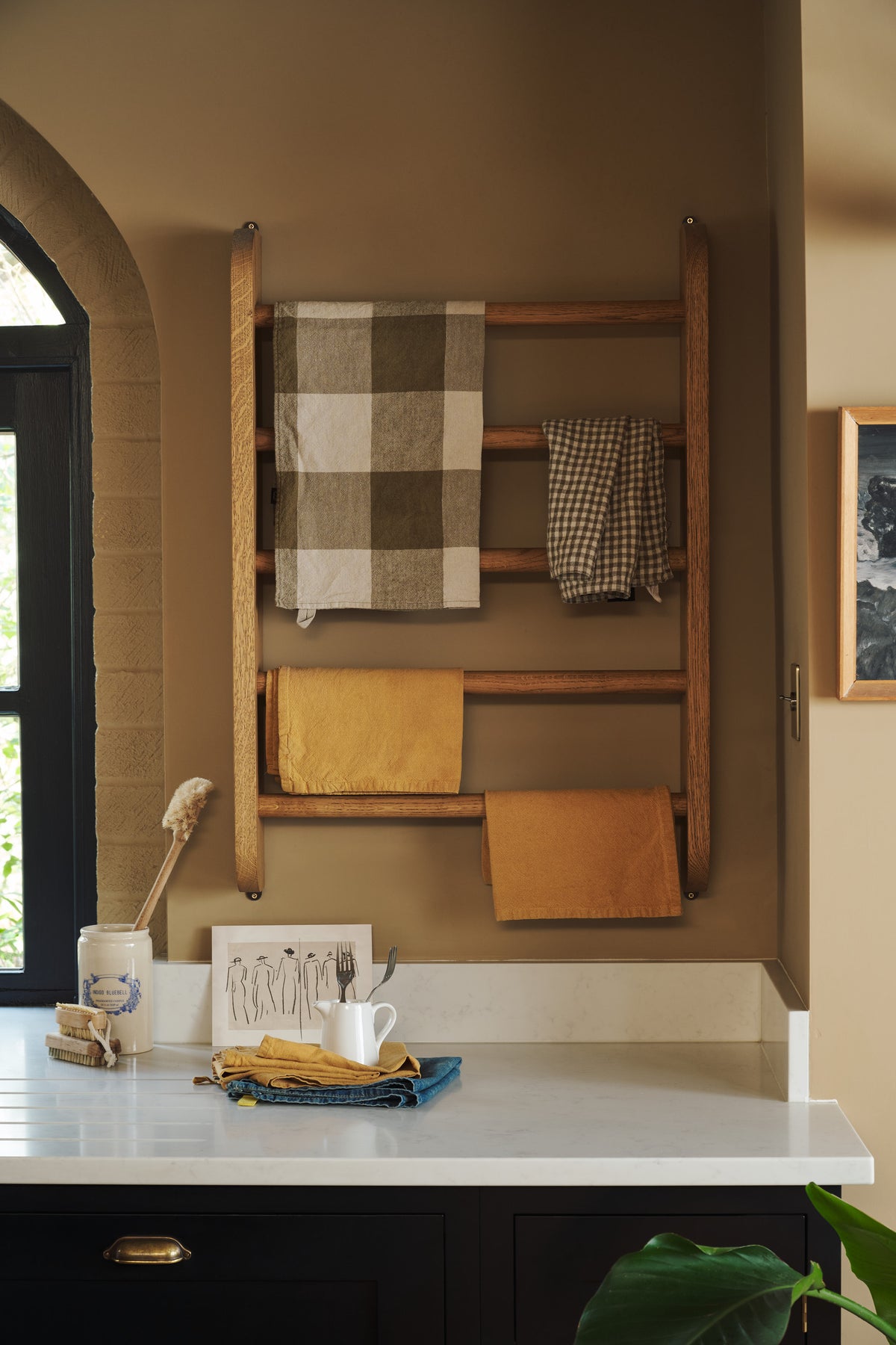 A close-up of a large, open Printer’s Black Shaker kitchen features a long cooker and sink run with a Silestone worktop, and a Haberdasher's wall hanging rack. Decorated with artisan ceramics and tea towels, the room feels carefully considered, comfortable and highly functional.