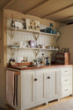 A close-up of a large, open Printer’s Black Shaker kitchen features a long kitchen island with a Silestone worktop, and both an open and closed pantry larder. Decorated with an aged brass Mayan tap, ceramics, dried produce, and baking equipments, the room feels carefully considered, comfortable and highly functional.