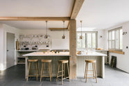 A tranquil and calm Shaker kitchen features a long sink and cooker run with a Silestone worktop and splashback, a single Belfast sink with a nickel Ionian tap, a long kitchen island with a breakfast nook, and shelves. Beautifully decorated with vintage stools that match the raw wooden beams, muted ceramics, silver pendant lights, and a steel firewood basket, this kitchen creates a lovely, simple space that embraces natural materials.