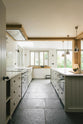 A tranquil and calm Shaker kitchen features a long sink and cooker run with a Silestone worktop and splashback, a single Belfast sink with a nickel Ionian tap, a long kitchen island with a breakfast nook, and shelves. Beautifully decorated with a chopping board that matches the wooden beams, some artisan ceramics, silver pendant lights, and an open book, this kitchen creates a lovely, simple space that embraces natural materials.