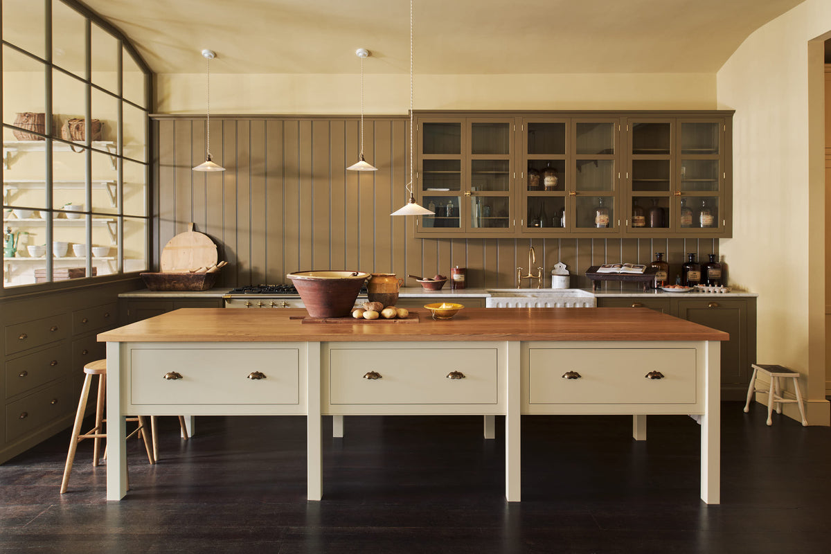 A simple Quaker cream and Folgate green Shaker kitchen features a cooker and sink run with Carrara marble worktop, glazed wall cupboards, and a bespoke prep table with a classic oak worktop. Lovingly adorned with milk pendant lights, artisan ceramics, antiques, and Helen stools, it creates a warm and inviting atmosphere.