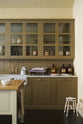 A quiet corner of a simple Quaker cream and Folgate green Shaker kitchen features a cooker and sink run with Carrara marble worktop, glazed wall cupboards, and a bespoke prep table with a classic oak worktop. Lovingly adorned with milk pendant lights, artisan ceramics, a fresh carton of eggs, and antique furniture, it creates a warm and inviting atmosphere.