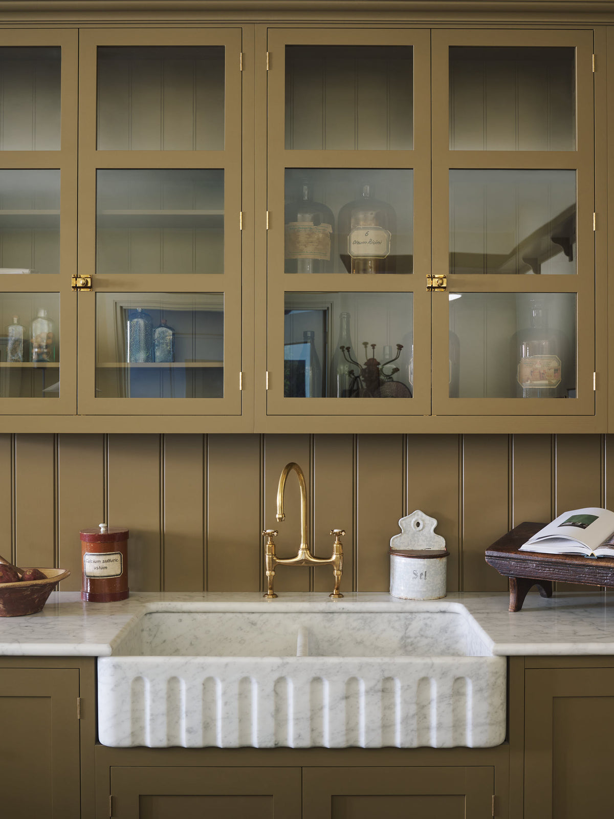 A close-up of a simple Quaker cream and Folgate green Shaker kitchen features a cooker and sink run with Carrara marble worktop, an aged brass Ionian tap, and glazed wall cupboards. Lovingly adorned with artisan ceramics, and antiques, it creates a warm and inviting atmosphere.