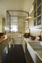 A simple Quaker cream and Folgate green Shaker kitchen features a cooker and sink run with Carrara marble worktop, glazed wall cupboards, and a bespoke prep table with a classic oak worktop. Lovingly adorned with milk pendant lights, artisan ceramics, antiques, and a Helen stool, it creates a warm and inviting atmosphere.