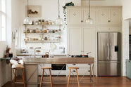A tall and simple mushroom-coloured kitchen features a Shaker cupboard run with a full-height cabinet, a large steel cooker and sink kitchen island, and shelves with hanging rails. Adorned with Helen stools, glass jars of dried ingredients, an amber glaze teapot, glass and steel pendant lights, a silver fridge, and still-life paintings, it creates a humble industrial kitchen space.