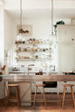 A tall and simple mushroom-coloured kitchen features a Shaker cupboard run with a full-height cabinet, a large steel cooker and sink kitchen island, and shelves with hanging rails. Adorned with Helen stools, glass jars of dried ingredients, an amber glaze teapot, glass and steel pendant lights, a silver fridge, and still-life paintings, it creates a humble industrial kitchen space.