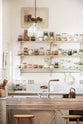 A closer look at a tall and simple mushroom-coloured kitchen features a Shaker cupboard run with a full-height cabinet, a large steel cooker and sink kitchen island, and shelves with hanging rails. Adorned with Helen stools, glass jars of dried ingredients, an amber glaze teapot, a glass and steel pendant light, and still-life paintings, it creates a humble industrial kitchen space.