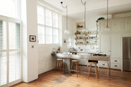 An oblique view of a tall and simple mushroom-coloured kitchen features a Shaker cupboard run with a full-height cabinet, a large steel cooker and sink kitchen island, and shelves with hanging rails. Adorned with Helen stools, glass jars of dried ingredients, an amber glaze teapot, glass and steel pendant lights, a silver fridge, and still-life paintings, it creates a humble industrial kitchen space.