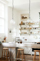 An oblique view of a tall and simple mushroom-coloured kitchen features a Shaker cupboard run with a full-height cabinet, a large steel cooker and sink kitchen island, and shelves with hanging rails. Adorned with Helen stools, glass jars of dried ingredients, an amber glaze teapot, glass and steel pendant lights, and still-life paintings, it creates a humble industrial kitchen space.