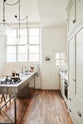Nestled between a Shaker cupboard run with a full-height cabinet and a large steel cooker and sink kitchen island, is a tall and simple mushroom-coloured kitchen. Adorned with Helen stools, glass jars of dried ingredients, an amber glaze teapot, a glass and steel pendant light, and still-life paintings, it creates a humble industrial kitchen space.