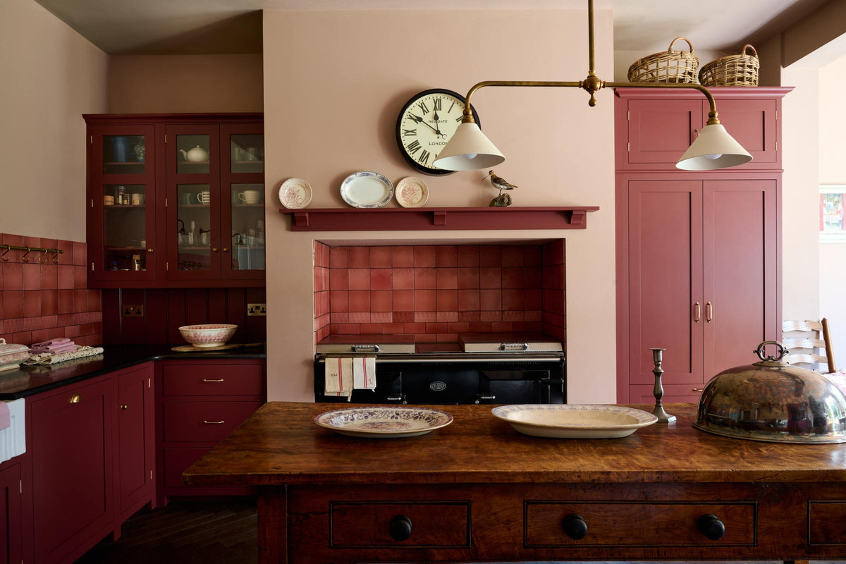 A humble Refectory red shaker kitchen featuring a sink run, glazed cupboards, a full-height pantry cupboard, and a large gas cooker in a chimney breast. The kitchen’s striking visuals are the Red Lace Market tiles used as a splashback in the chimney breast and along the sink run, enhancing the room's warmth. The space is decorated with vintage furniture, particularly an old, solid oak prep table.