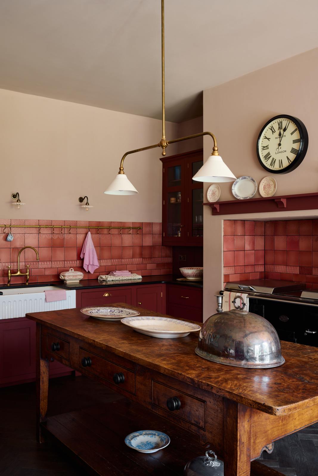 An angled view of a humble Refectory red shaker kitchen featuring a sink run, glazed cupboards, and a large gas cooker in a chimney breast. The kitchen’s striking visuals are the Red Lace Market tiles used as a splashback in the chimney breast and along the sink run, enhancing the room's warmth. The space is decorated with vintage furniture, particularly an old, solid oak prep table.