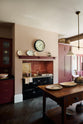 An angled view of a humble Refectory red shaker kitchen featuring a sink run, glazed cupboards,  a full-height pantry cupboard, and a large gas cooker in a chimney breast. The kitchen’s striking visuals are the Red Lace Market tiles used as a splashback in the chimney breast and along the sink run, enhancing the room's warmth. The space is decorated with vintage furniture, particularly an old, solid oak prep table.