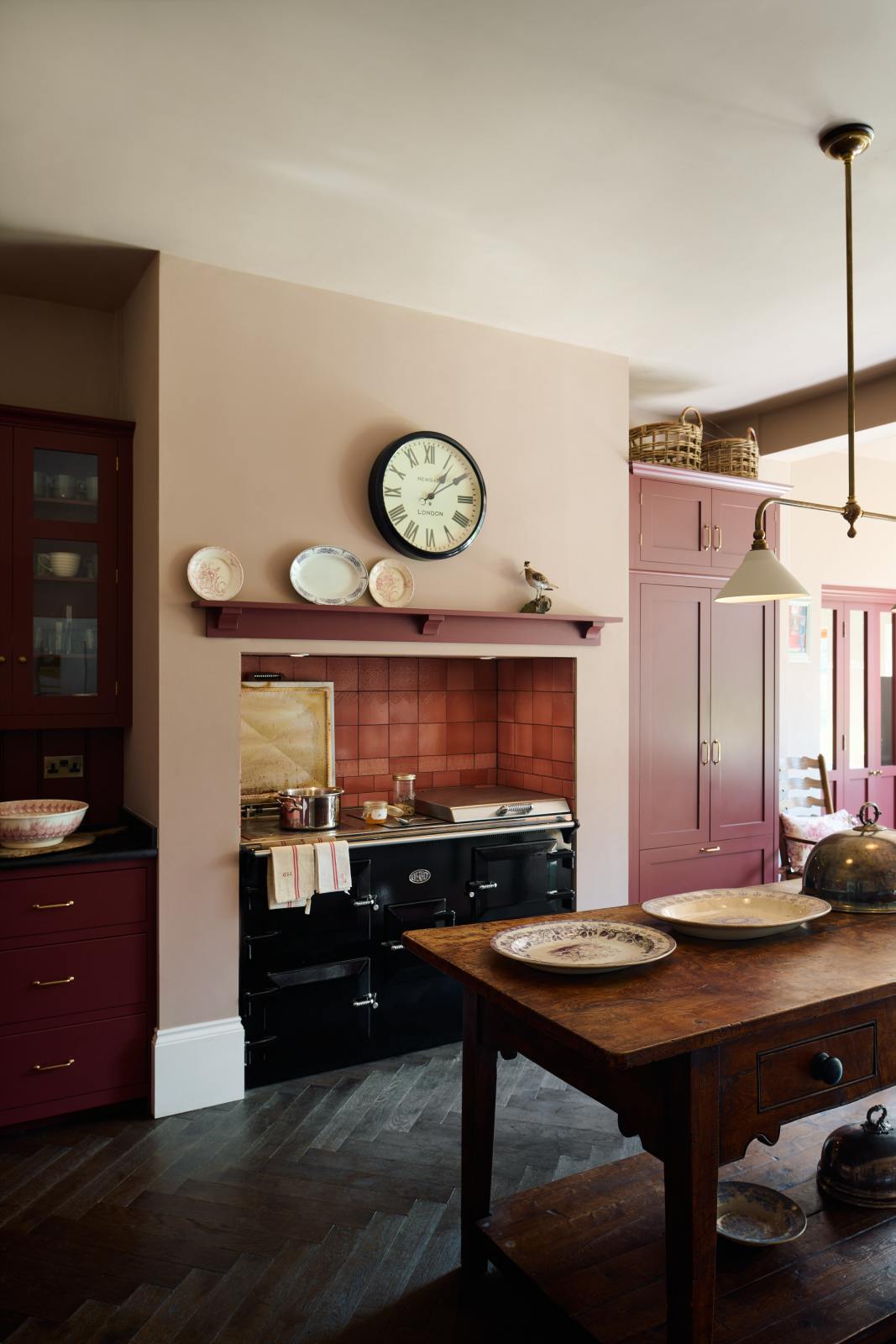 An angled view of a humble Refectory red shaker kitchen featuring a sink run, glazed cupboards,  a full-height pantry cupboard, and a large gas cooker in a chimney breast. The kitchen’s striking visuals are the Red Lace Market tiles used as a splashback in the chimney breast and along the sink run, enhancing the room's warmth. The space is decorated with vintage furniture, particularly an old, solid oak prep table.