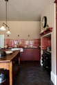 Looking into the sink run of a humble Refectory red shaker kitchen featuring glazed cupboards, a full-height pantry cupboard, a full-height cupboard and a large gas cooker in a chimney breast. The kitchen’s striking visuals are the Red Lace Market tiles used as a splashback in the chimney breast and along the sink run, enhancing the room's warmth. The space is decorated with vintage furniture, particularly an old, solid oak prep table.