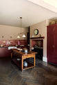 An angled view of a humble Refectory red shaker kitchen featuring a sink run, glazed cupboards,  a full-height pantry cupboard, and a large gas cooker in a chimney breast. The kitchen’s striking visuals are the Red Lace Market tiles used as a splashback in the chimney breast and along the sink run, enhancing the room's warmth. The space is decorated with vintage furniture, particularly an old, solid oak prep table and beautiful rich oak parquet floors.