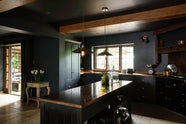 An angled view of the sophisticated Pantry Blue shaker kitchen with a cooker and sink run, along with a vintage island topped with Iroko wood. Light stone flooring enhances the space, featuring wooden ceiling accents, a large cooker splashback, and two hanging lights above the island.