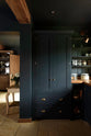 A closer look at the tall Pantry Blue double-door pantry cupboard, featuring open shelving and a cabinet with an Iroko wood worktop. The light stone flooring enhances the space, complemented by wooden ceiling accents.
